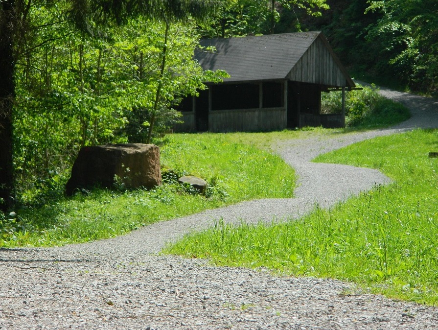 Schmaler, gewundener Kiesweg, der durch eine grüne Waldlichtung zu einer einfachen hölzernen Schutzhütte mit Satteldach führt.